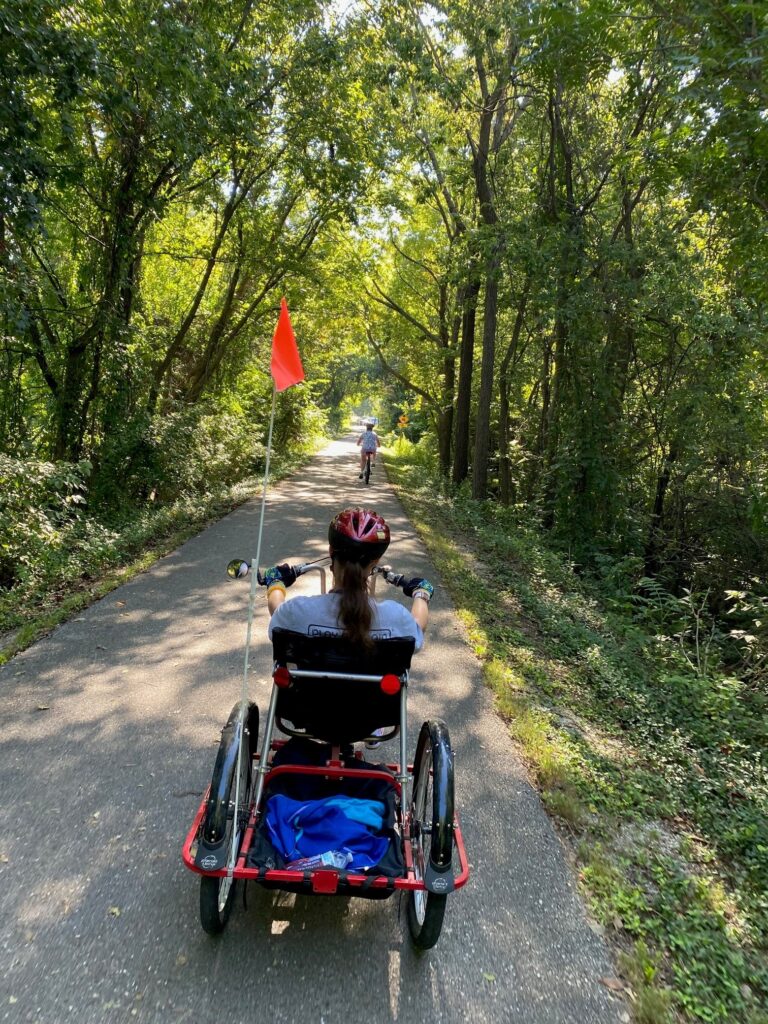 Picture shows a person riding an accessible bicycle on trail.