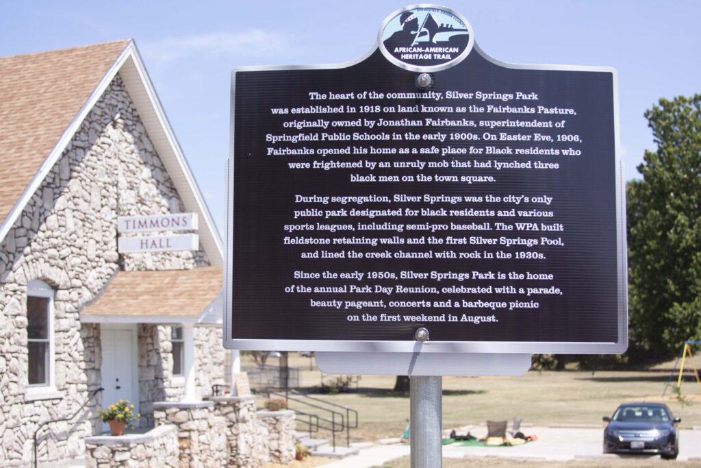 Picture of the African-American Heritage Trail sign that is posted by Timmons Hall in Silver Springs Park. The trail follows  the Jordan Creek trail through the park in Springfield MO). The sign reads: "The heart of the community, Silver Springs Park was established in 1918 on land known as the Fairbanks Pasture, originally owned by Jonathan Fairbanks, superintendent  of Springfield Public Schools in the early 1900s. On Easter Eve, 1906, Fairbanks opened his home as a safe place for Black residents who were frightened by an unruly mob that had lynched three black men on the town square.
During segregation, Silver Springs was the city's only public park designated for black residents and various sports leagues, including semi-pro baseball. The WPA built fieldstone retaining walls and the first Silver Springs Pool, and lined the creek channel with rock in the 1930s. 
Since the early 1950s, Silver Springs Park is the home of the annual Park Day Reunion, celebrated with a parade, beauty pageant, concerts and a barbeque picnic on the first weekend in August."